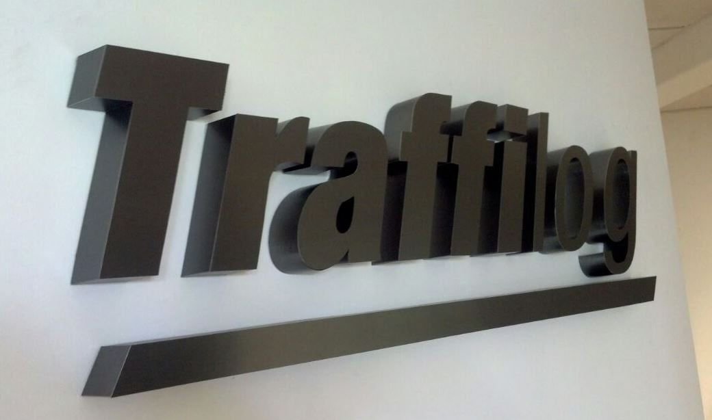 Letras 3D de aluminio anodizado y cepillado color grafito con el logotipo de Traffilog sobre muro blanco